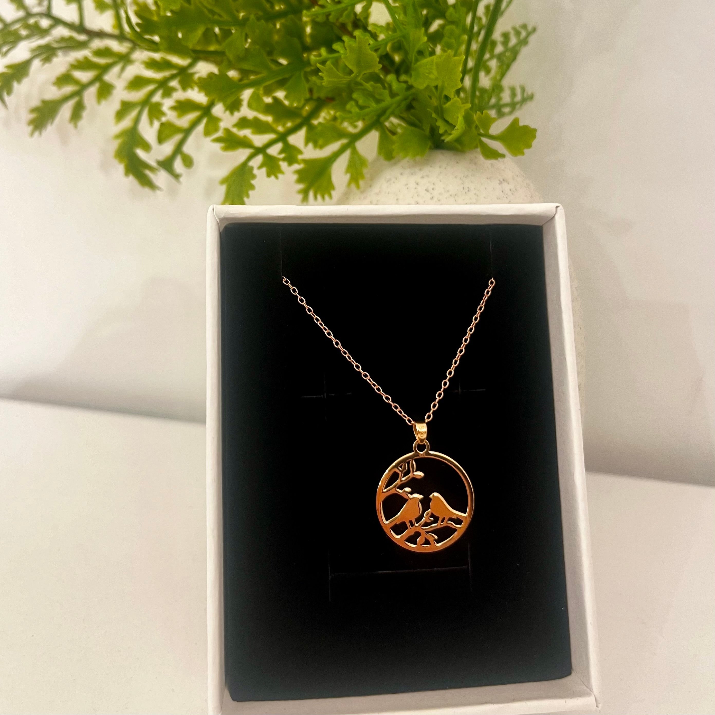 Gold necklace with a circular pendant featuring two birds in flight, displayed in a box with green foliage in the background.